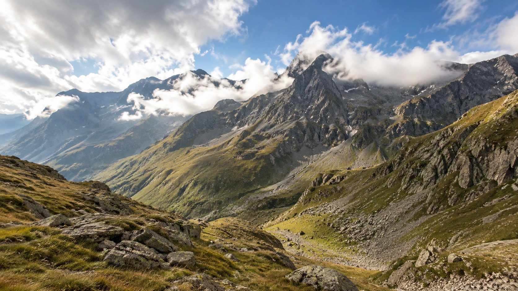 Weitgehend schneefreie Schweizer Hochalpen mit Fels, Geröll und Gras unter aufziehenden Wolken.