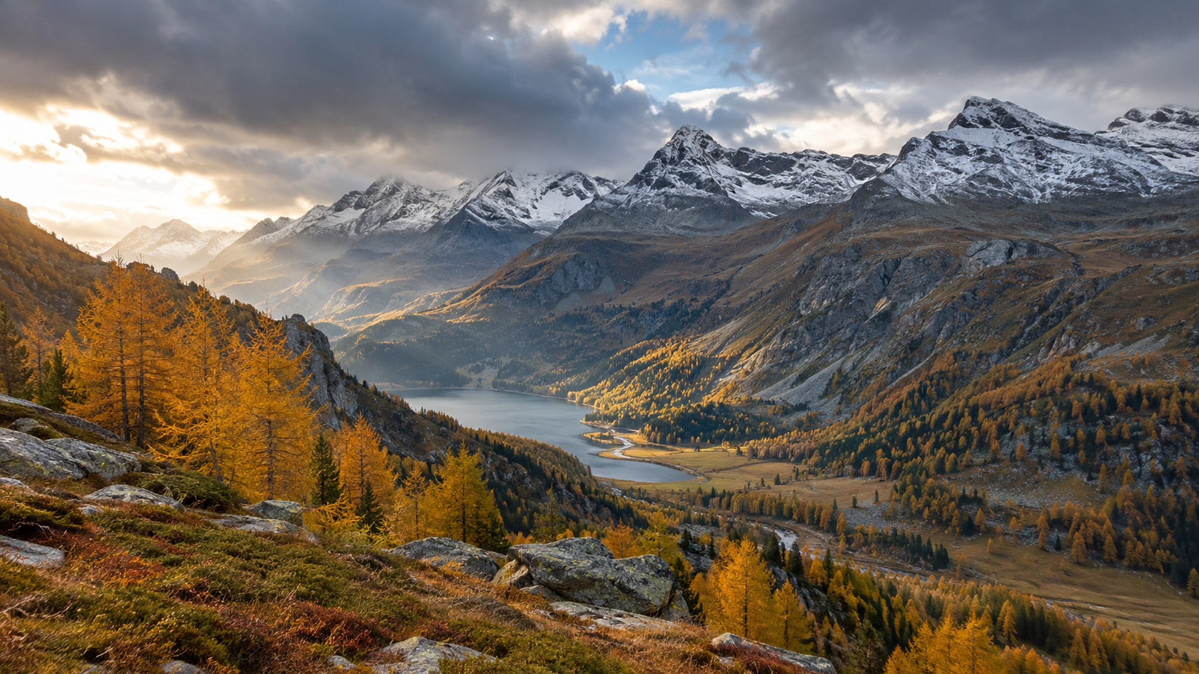 Herbstliche Schweizer Alpen mit golden verfärbten Lärchen, einem Bergsee im Tal und frisch angezuckerten Gipfeln unter dramatischen Wolken.